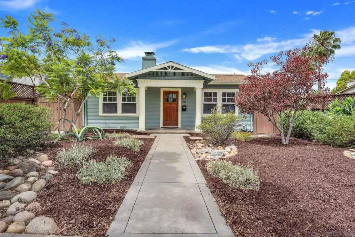 Charming single-story craftsman bungalow with sage green horizontal siding, white trim, and brown front door, featuring drought-tolerant landscaping with native plants, ornamental grasses, and a concrete walkway leading to the entrance
