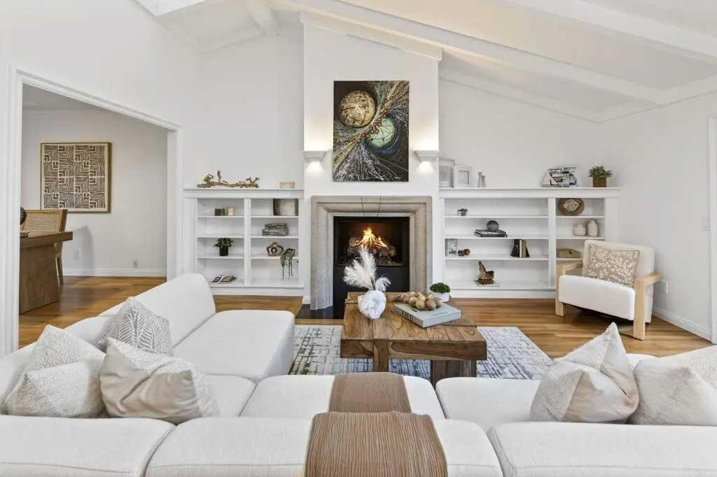Bright living room with white sectional sofa, rustic wood coffee table, fireplace with white built-in shelving on both sides, hardwood floors, and contemporary artwork above the mantel